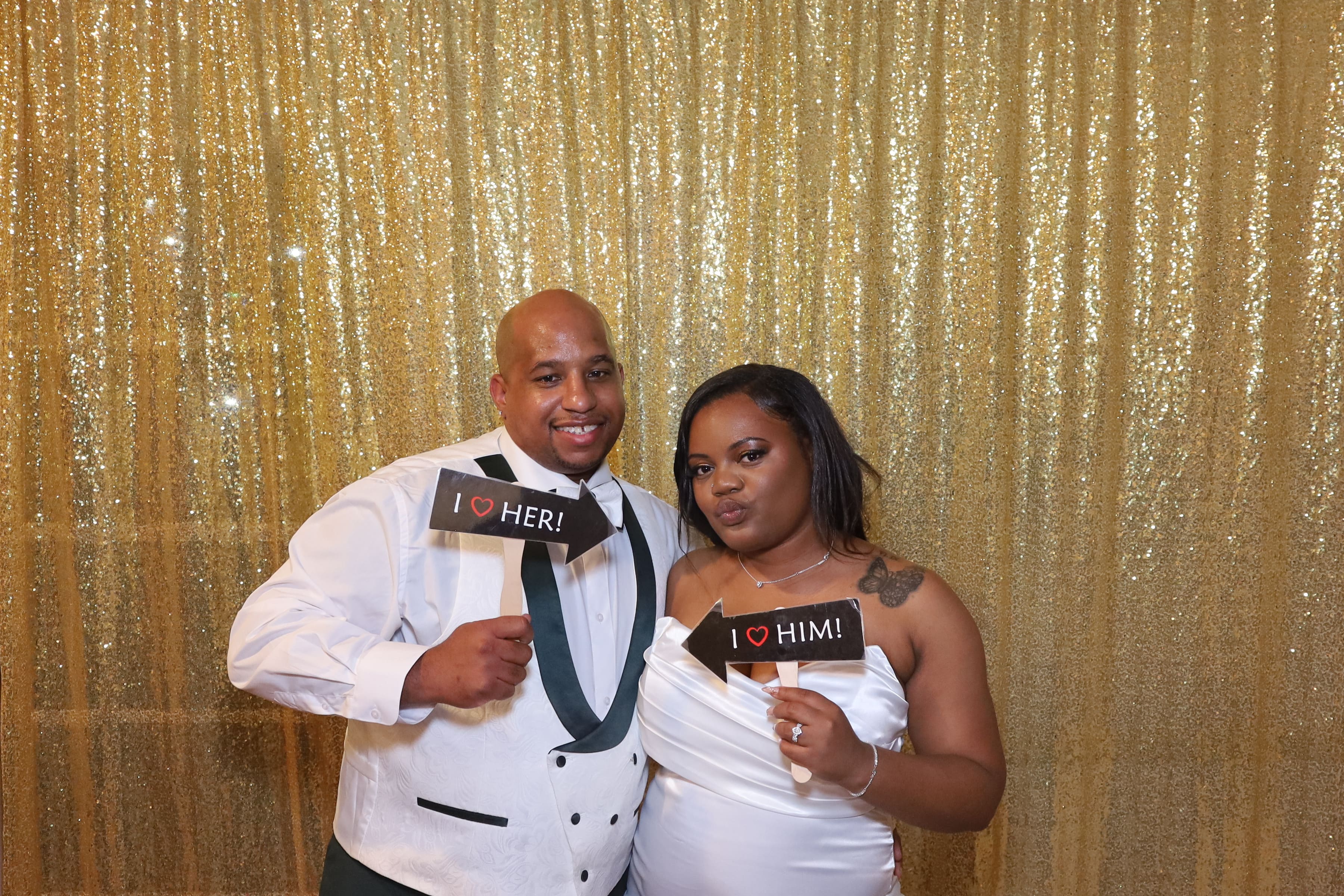Wedding couple with 'I HEART HIM' and 'I HEART HER' signs, gold sequin backdrop